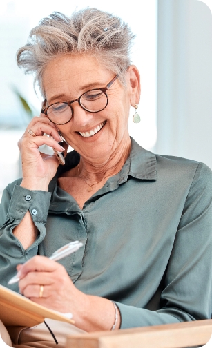 Newcastle Specialist Prosthodontics Older woman smiling while talking on the phone and writing in a notebook.